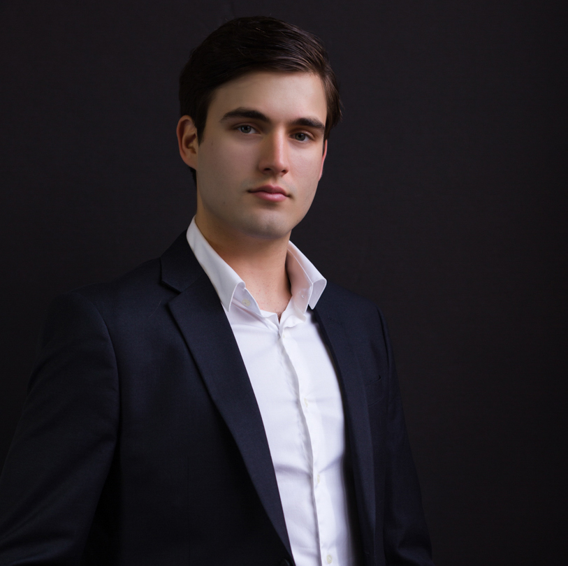 Business headshot on dark background of young male executive in jacket and white dress shirt with open collar.