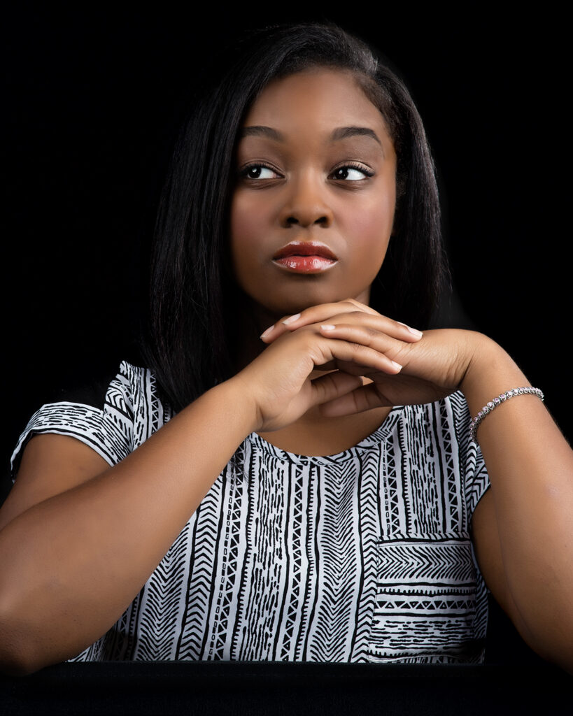 A pretty young black musical theater actress poses for a dramatic headshot against a dark background.