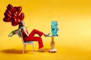 Feature image for fashion shoe designer's Valentines Day ad campaign, “Celebrate Love”, by Houston fashion photographer Gerard Harrison. Against a graduated, bright yellow background. a beautiful, happy young woman in red jumpsuit sits in a white antique, wooden chair with one elegantly shod foot on a the edge of the white antique table in front of her. She holds a windblown bouquet of heart balloons, and the brightly colored silk scarf around her neck, blows in the breeze behind her. A single rose and stack of the designer’s signature turquoise shoe boxes sits on the table in front of her. An empty space at the right creates space for ad text.