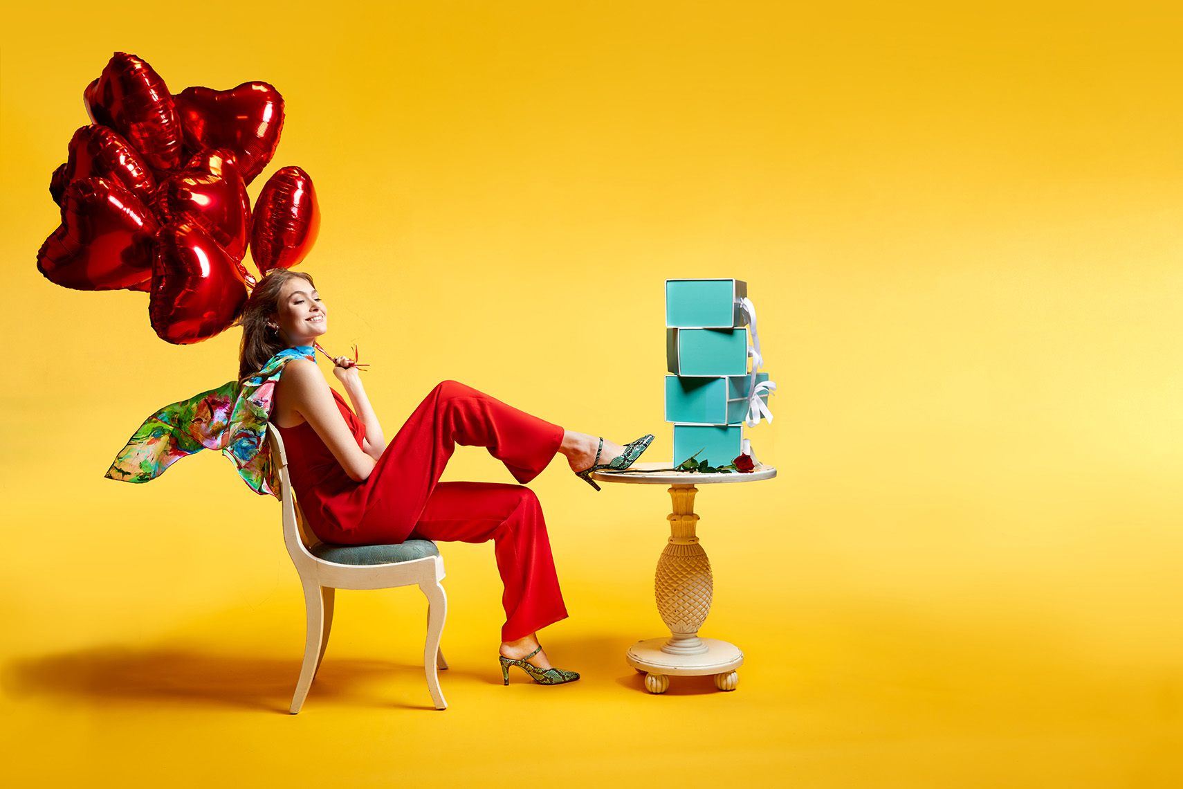 fashion-photo-shoe-designer-campaign-Gerard-Harrison-Photographer-Houston Shoe designer ad for Valentines Day campaing, featuring woman in red jumpsuit, sitting at a table holding windblown bouquet of heard balloons, and stack of turquoise shoe boxes in front of her, against a graduated yellow background.