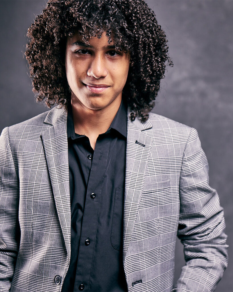 Headshot for young actor with long, curly hair, wearing plaid sport coat, dark, open collar shirt.