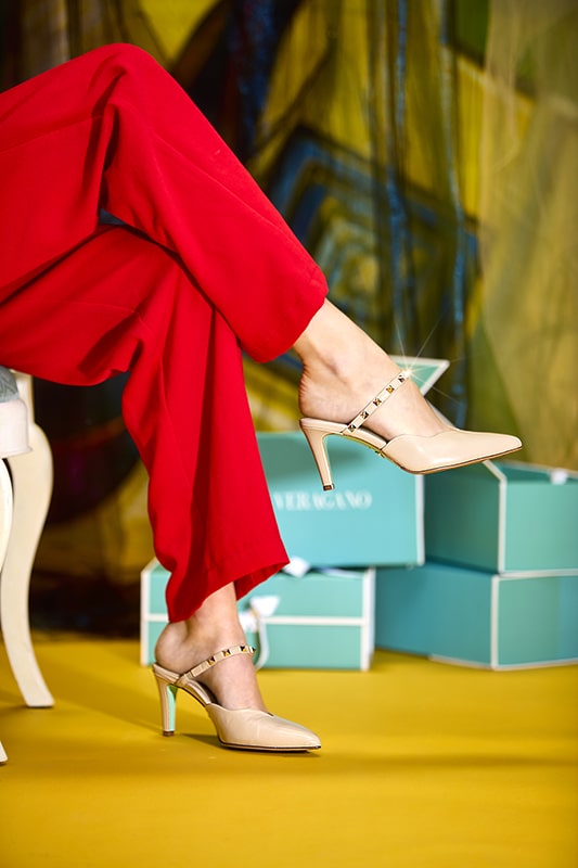 A model crosses her legs, showing a stylish pair of rhinestone adorned nude pumps. A stack of turquoise designer shoe boxes sits on the floor in the background, and behind them is a glimpse of Veragano’s signature surrealist painting.