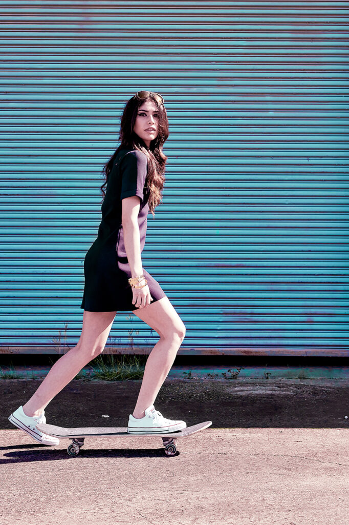 Fashion advertising photo of a female model in casual to dressy mini dress and sneakers rides a skateboard past a colorful industrial building.