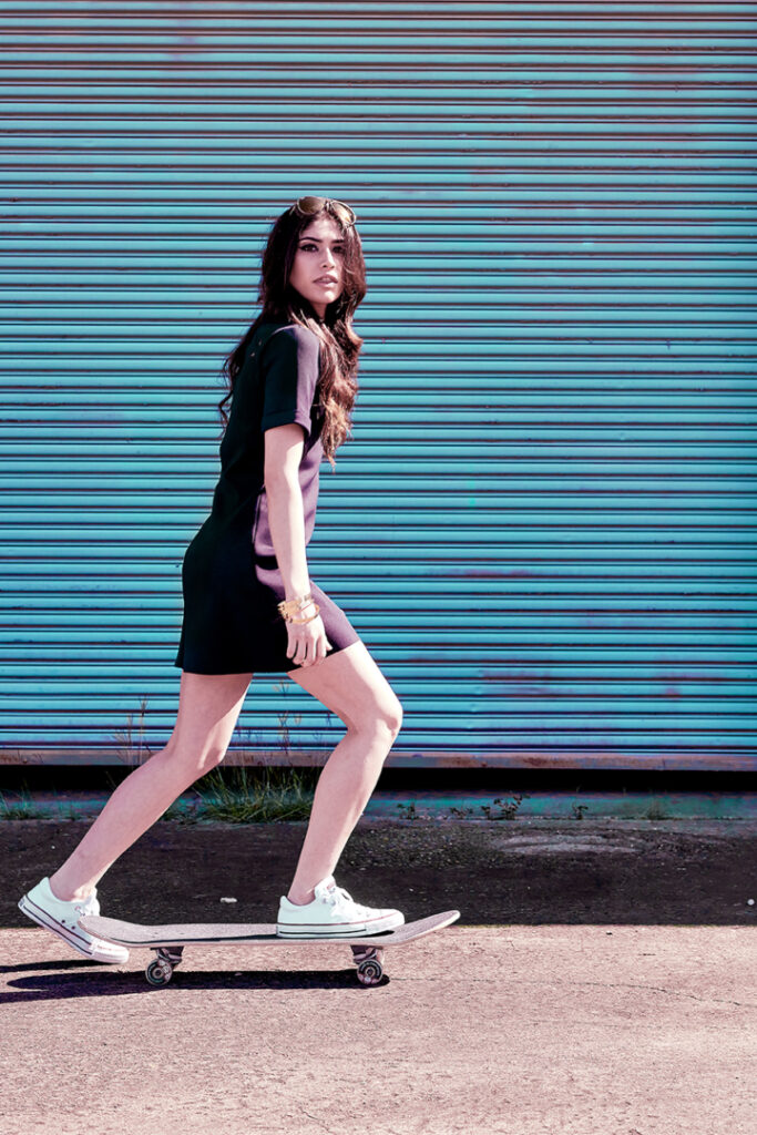 A pretty young woman in a stylish minidress and sneakers rides a skateboard in front of a turquoise industrial garage door.