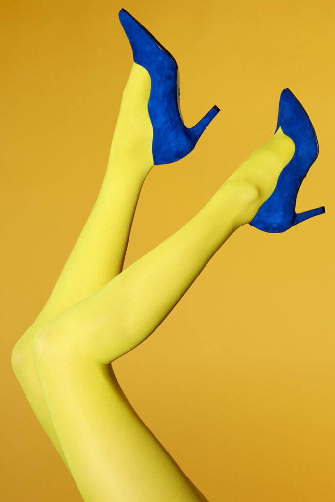 Legs in yellow stocking with blue pumps held in the air against a bright orange-yellow background.