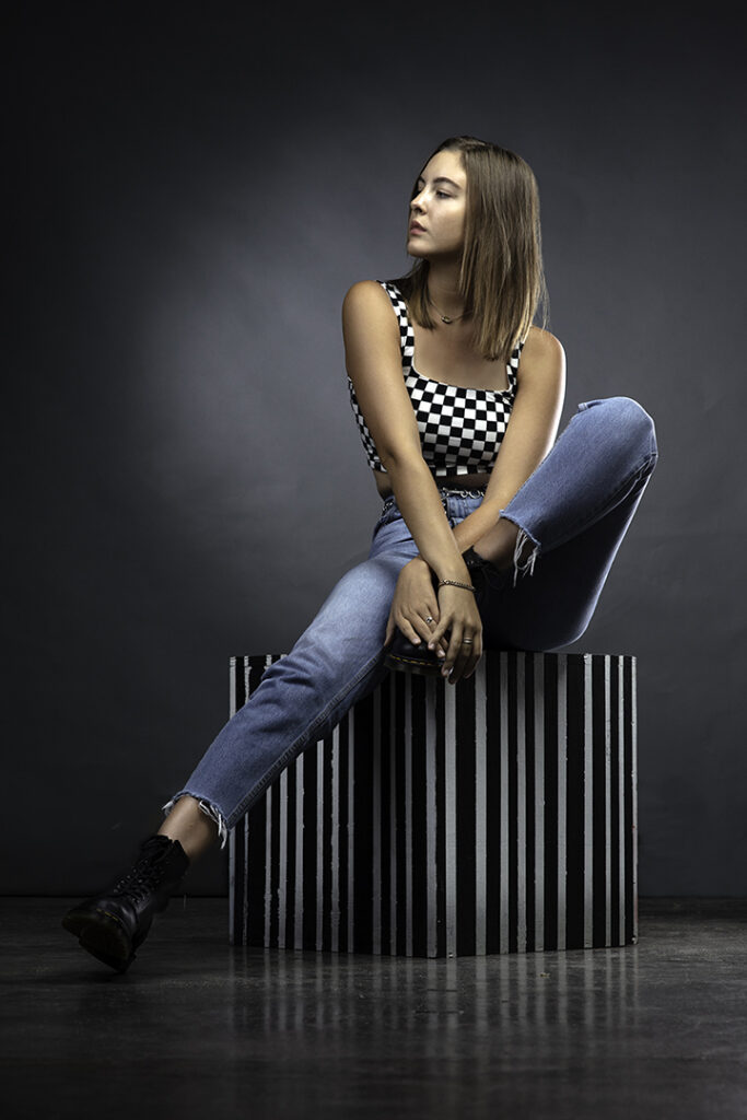 Beautiful female model poses on black and white striped cube for modeling portfolio in jeans and checkerboard halter top.