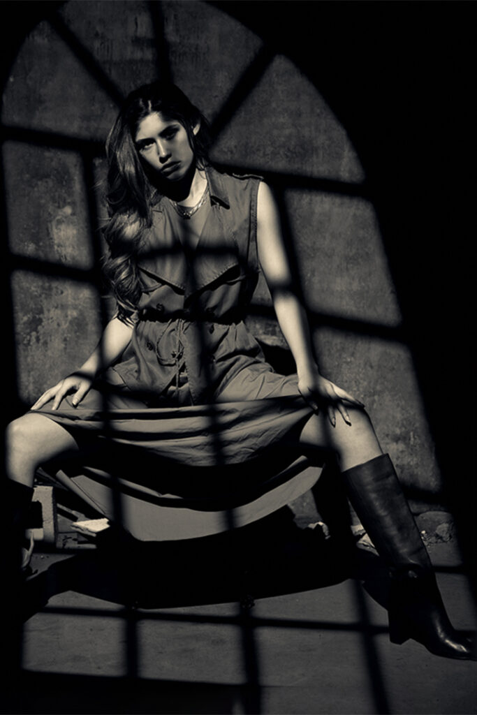 Sepia toned black and white photo of model in dark setting, framed by window pane shadow.
