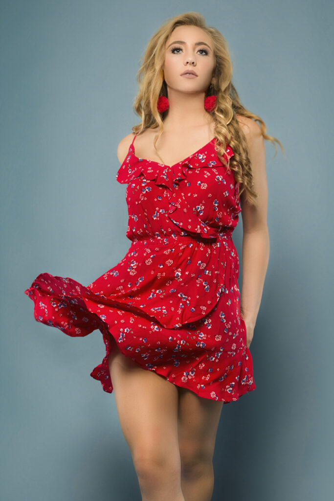 Beautiful blonde model in short red sundress appears to dance as skirt flutters above her legs.