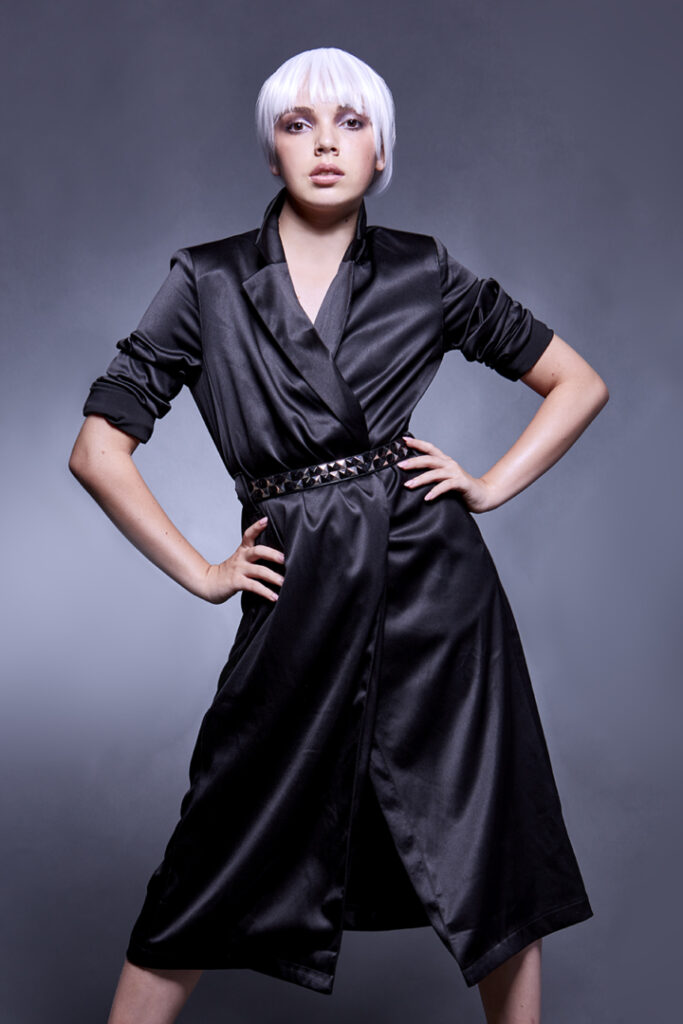 Model with white hair strikes angular hands on hips pose in black satin dress for modeling portfolio.