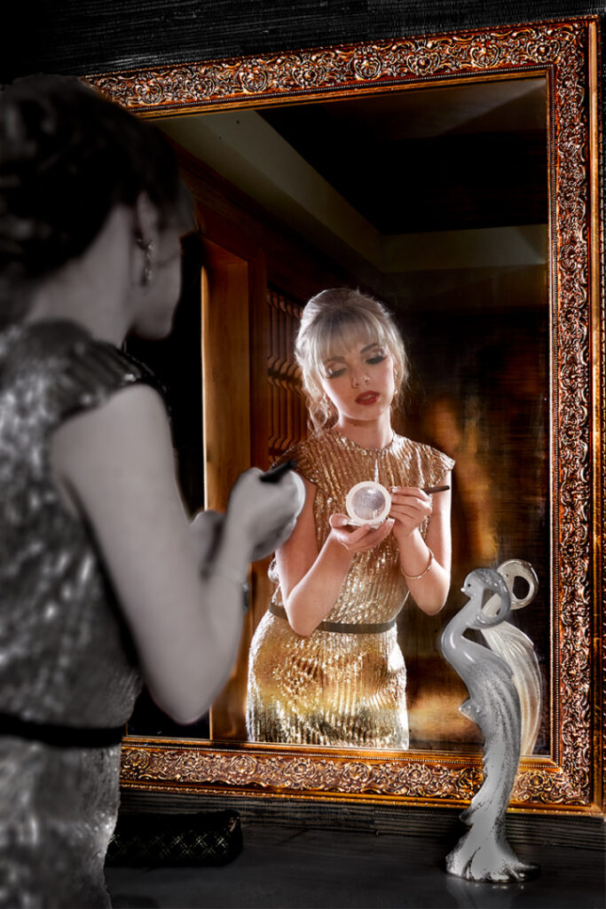 A model partly in shadow, applies makeup before a mirror that glows with an unnatural brightness. Part of a fashion editorial.