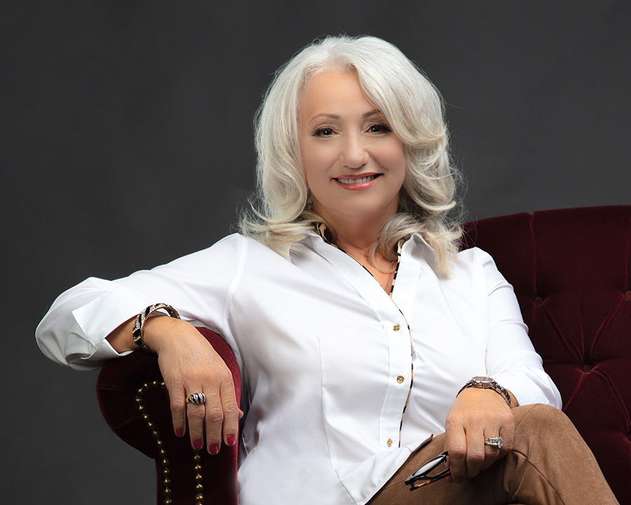 Casual headshot for senior female business owner with white hair relaxing on a sofa. Dark gray background.