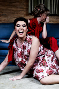 Fashion editorial photo of two models laughing. One is seated on a vintage sofa and the other in front of her, on the floor. Fashion editorial by Gerard Harrison, Houston photographer.