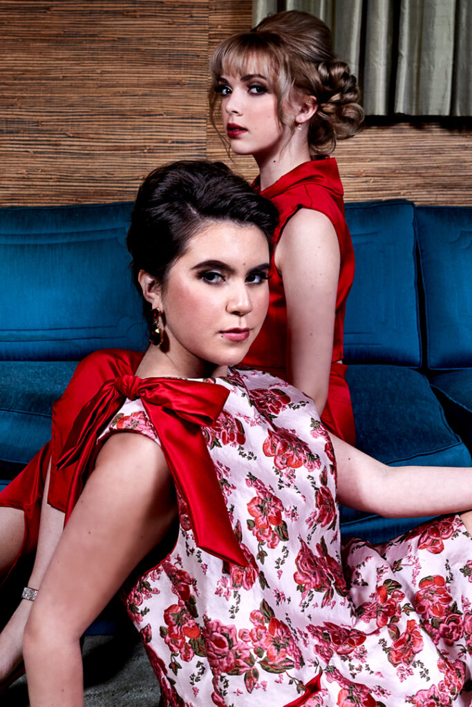 Fashion editorial photo of two models wearing serious expressions. One is seated on a vintage sofa and the other in front of her, on the floor. Fashion editorial by Gerard Harrison, Houston photographer.
