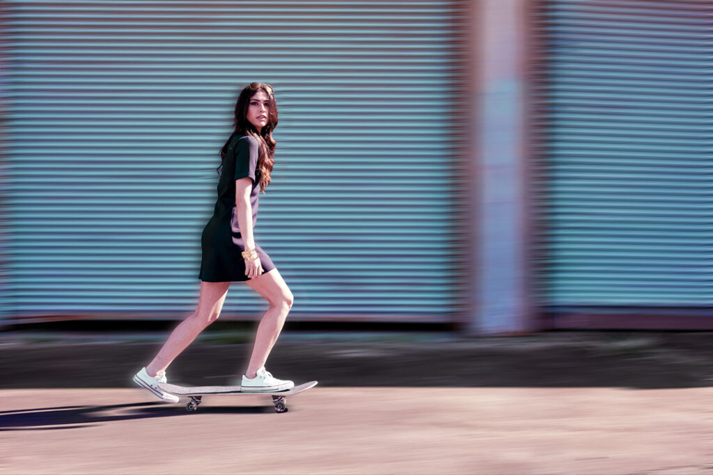 Young woman in little black dress and sneakers rides a skateboard to the office in East Downtown Houston. Dress by Work in Process Style. Gerard Harrison Houston fashion photographer.