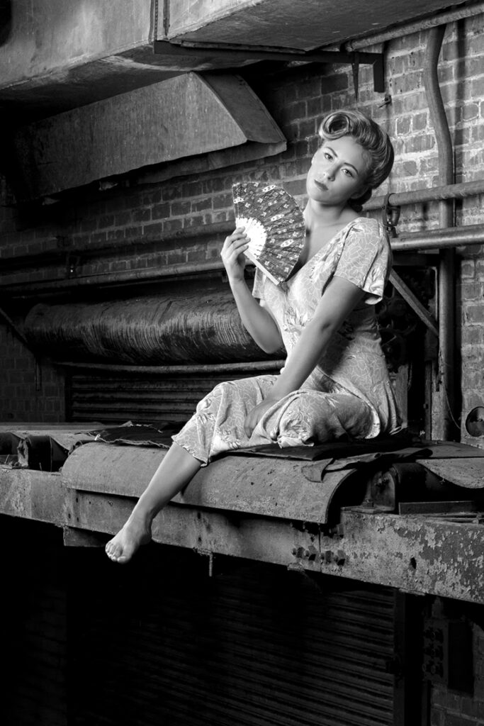 Vintage fashion editorial image of young woman in a 1940s silk print dress, seated on the rusty conveyor in the abandoned Imperial Sugar plant, Sugar Land, Texas. Model Brenna Smith. Gerard Harrison, Houston fashion photographer.